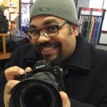 Melqui Rodriguez is wearing a knit hat and glasses. He is holding a camera with the lens pointed toward the camera (viewer of the photograph).