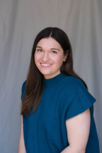 This is a woman, Jennifer Crim, with long straight dark hair. She is wearing a turquoise top and smiling in front of a gray backdrop.